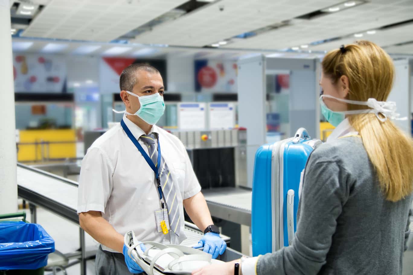 Travelers placing their belongings into trays at an airport security checkpoint, with X-ray machines and TSA officers in the background